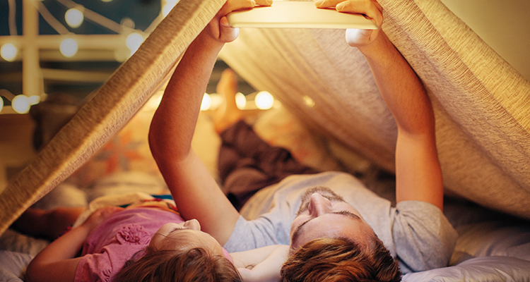 Vater und Kind liegen im Zelt und schauen etwas auf einem Tablet an