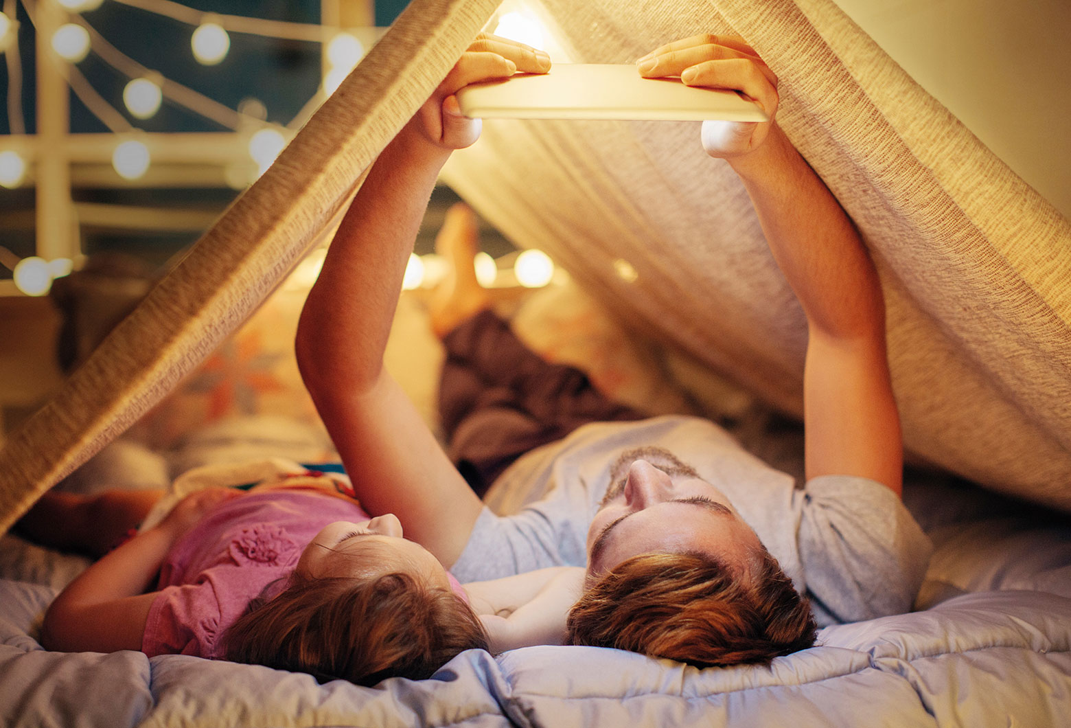 Vater und Kind liegen im Zelt und schauen etwas auf einem Tablet an