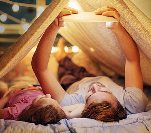 Vater und Kind liegen im Zelt und schauen etwas auf einem Tablet an